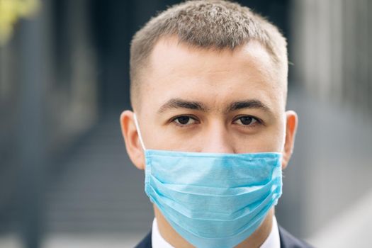 Close up of face of young handsome businessman in medical mask looking straight to camera with happy look. Portrait of man at street. Coronavirus concept.