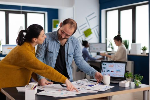 Woman in start up office explaining charts to executive businessman pointing at charts. Diverse team of business people analyzing company financial reports from computer.