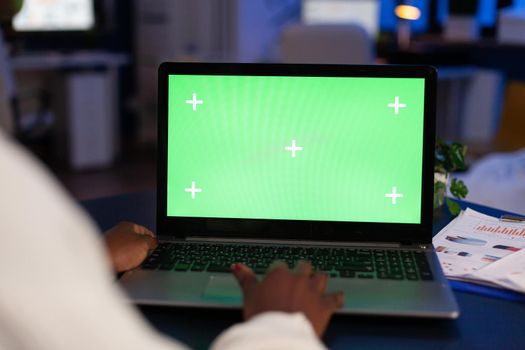 Black freelancer working on laptop with green screen display, green mockup, chroma key desktop sitting at desk in business office working at night. Freelancer watching at isolated desktop overwork.