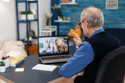 Old parent greenting daughter during video conference on portable computer. Happy grandfather saying hello in the course of online video confencere with family from living room.