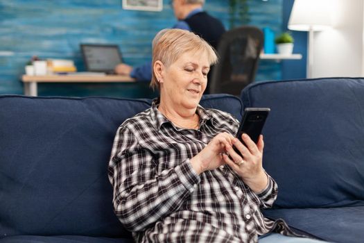 Hapy elderly lady texting on phone relaxing on sofa enjoying retirement lifestyle. Senior woman browsing on internet using smartphone sitting on sofa in home living room.