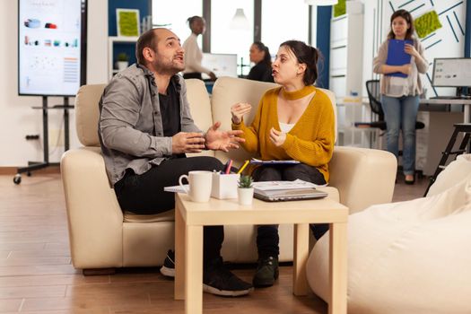 Business colleagues arguing, yealing each other during work hours sitting on the couch, while diverse colleagues working scared in background. Coworkers having conflict because of mistakes from project