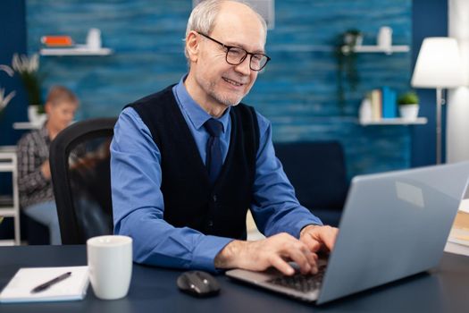 Smart senior businessman working on laptop wearing a tie and glasses. Elderly man entrepreneur in home workplace using portable computer sitting at desk while wife is holding tv remote.