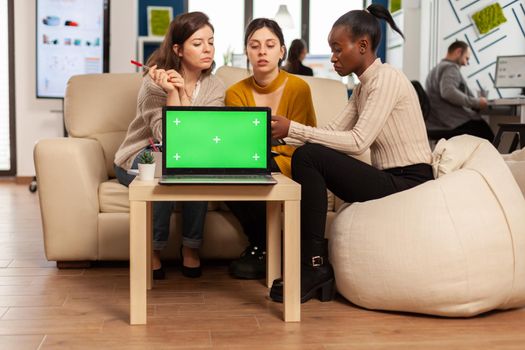 Laptop with green screen on table in start up company with multiethnic diverse coworkers working together behind it. African american woman discussing project strategy with colleques looking at tablet pc.