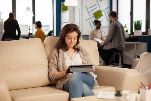 Employee taking break sitting on confortable couch holding tablet browsing, reading great news and smiling. Multiethnic creative coworkers planning new financial project working in open plan office