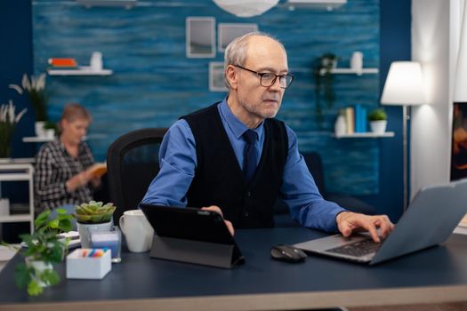 Senior manager working on presentation using laptop and tablet pc sitting at office. Elderly man entrepreneur in home workplace using portable computer sitting at desk while wife is reading a book sitting on sofa.