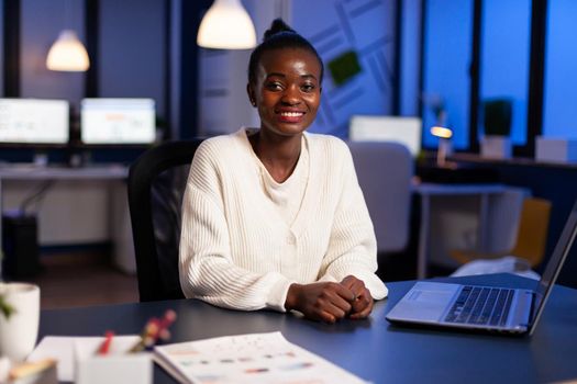 Close up portrait of business african woman smiling at camera working using laptop sitting in startup company late at night. Focused employee doing overtime respecting deadline of project.