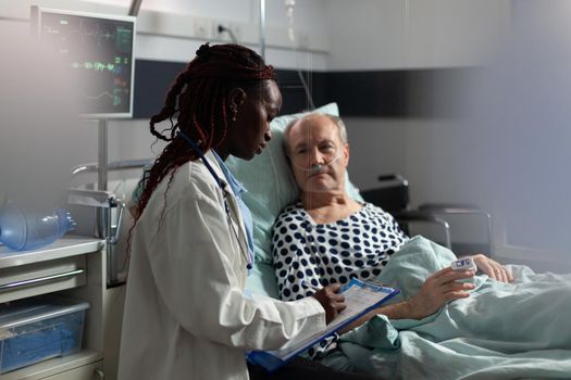 African american doctor in hospital room discussing diagnosis and treatment with sick senior man laying in bed reassuring full recovery. Unwell patient breathing through oxygen mask.