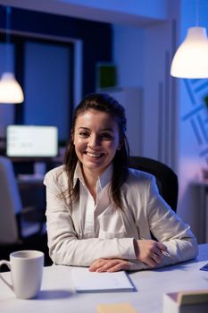 Close up portrait of business woman smiling at camera after drinking cup of coffee sitting at desk in business office late at night. Focused employee using technology network wireless for financial statistics