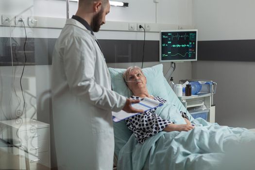 Sick senior woman lying in hospital bed listening doctor talking about treatment and diagnosis. Patient inhale and exhale with help from oxygen tube. Iv drip bag attached on arm.