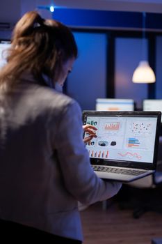 Back view of manager woman analysing financial statistics holding laptop standing in start-up business office late at night overworking. Tired focused employee using modern technology network wireless