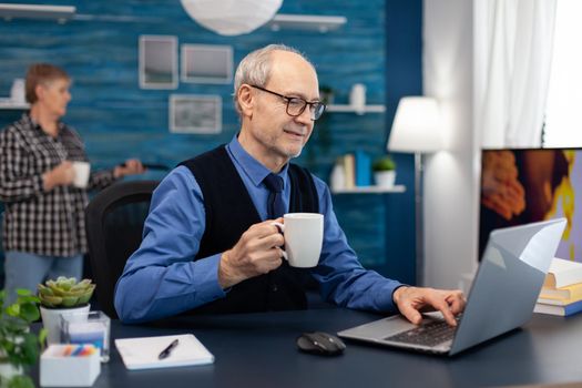 Senior businessman holding cup of coffee working on laptop. Elderly man entrepreneur in home workplace using portable computer sitting at desk while wife is holding tv remote.