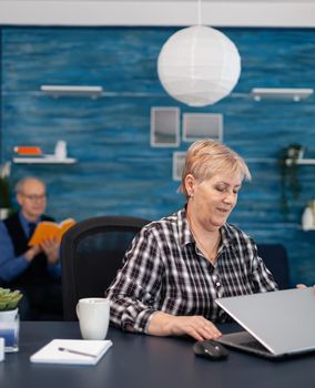 Mature entrepreneur sitting in front of portable computer in office. Elderly woman in home living room using moder technoloy laptop for communication sitting at desk indoors.