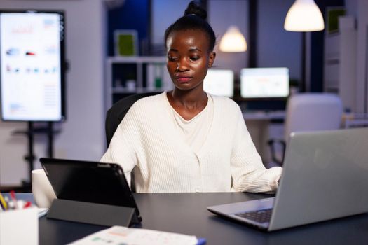 African businesswoman working late at night in office using tablet pc. Busy multitasking employee analysing financial statistics overworking writing, searching.