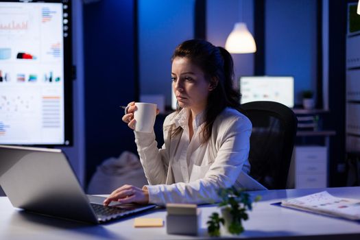 Freelancer woman with a cup of coffee tapping on business computer overworking at marketing project. Busy entrepreneur using modern technology network wireless late at night in company office