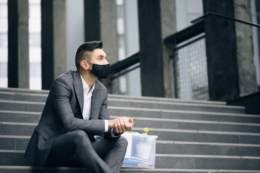 Unemployed businessman lost his business. Anxious concept. Workless man in despair. Fired male office worker in medical mask sitting on stairs in depression with box of stuff.