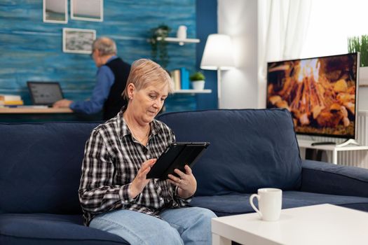 Senior lady scrolling on digital device relaxing on sofa. Elderly woman using moder technoloy tablet pc in home living room and husband working on laptop computer