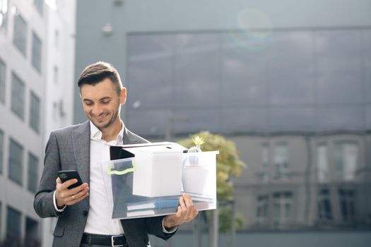 Happy businessman stand smiling use phone near business center. Businessman with box of personal stuff uses phone texting scrolling tapping. Portrait suit career male office handsome technology.