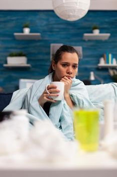 Woman with flu sitting at home drinking hot tea feeling sick. Person with temperature taking medicine for health virus infection, adult with disease pain headache
