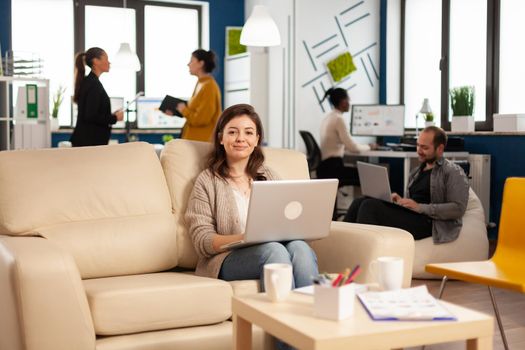 Portrait of entrepreneur typing on laptop looking at camera smiling while diverse team working in background. Multiethnic coworkers talking about startup financial company in modern business office