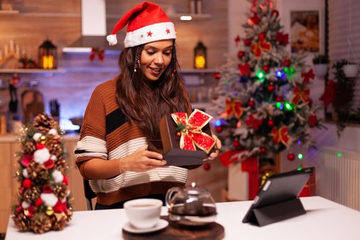 Caucasian woman opening gift box while using video call technology with friends on tablet. Festive young adult with sweater receiving present feeling joyful in christmas decorated kitchen