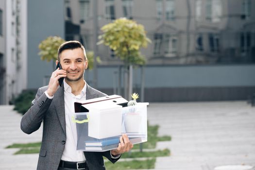 Cellphone conversation. An employee between office buildings with a box and documents with a desk flower. Happy fired man talking on phone with stuff in box. Speaking on telephone.