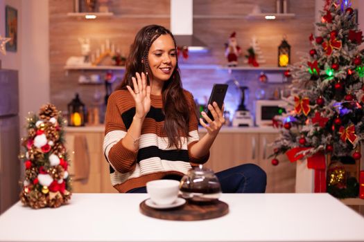 Happy woman holding smartphone for video call chatting conference on online internet with relatives in kitchen decorated with ornaments, lights and christmas tree for winter celebration