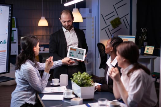 Entrepreneur businessman showing company strategy using tablet for corporate presentation working overtime in meeting office room late at night. Diverse multi-ethnic teamwork brainstorming ideas.