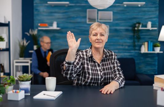 Cheerful elderly pensior waving at camera and husband is reading a book sitting on sofa. Happy senior woman with gray hair saying hello at camera during video call in home living room.