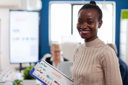 African woman manager looking at camera smiling, holding clipboard, while diverse coworkers talking in background. Manager working in professional start up financial business, modern company workplace ready for meeting.