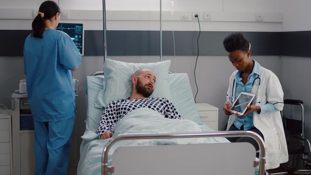 Practitioner black doctor explaining bones radiography to sick man using tablet computer in hospital ward. Patient with nasal oxygen tube lying in bed during disease recovery