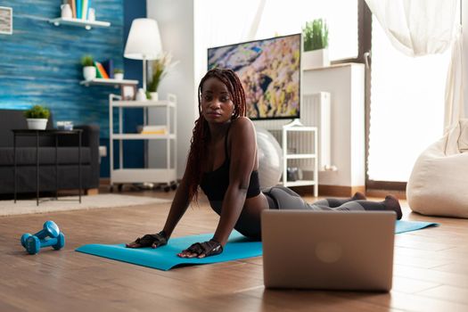 Black woman exercising and stretching her body doing flexibility exercises sitting on yoga mat in home living room wearing leggings during online tutorial. Workout and healthy lifestyle.