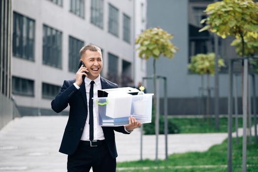 Happy fired man talking on phone with stuff in box. Cellphone conversation. Speaking on telephone. An employee between office buildings with a box and documents with a desk flower