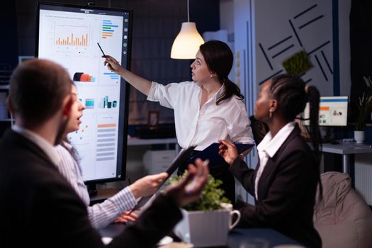Multi ethnic businesspeople brainstorming company ideas checking graphs statistics using tv standing at conference table. Diverse teamwork working at management presentation in meeting room