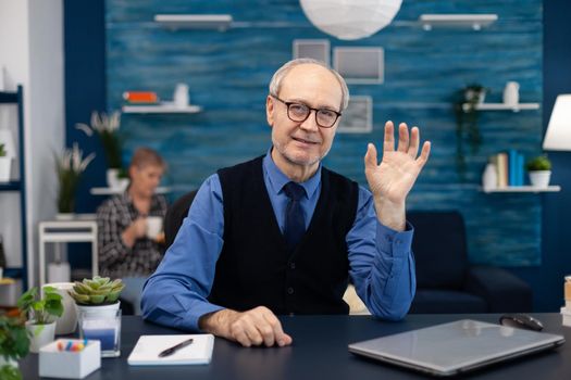 Senior businessman waving at camera wearing glasses during video call. Elderly man in the course of online conference waving at camera from home office sitting at desk wearing glasses.