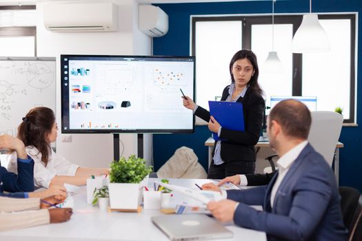 Young team leader in big corporation briefing coworkers pointing at graph meeting. Corporate staff discussing new business application with colleagues looking at screen