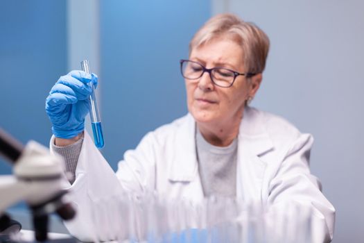 Scientist senior woman looking into test tube for biochemistry test. Doctor in white coat research a new experiment in laboratory, analyzing biotechnology work with modern equipment.