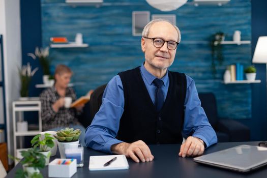 Adult man with gray hair looking pensive at camera wearing glasses. Elderly man entrepreneur in home workplace using portable computer sitting at desk while wife is reading a book sitting on sofa.