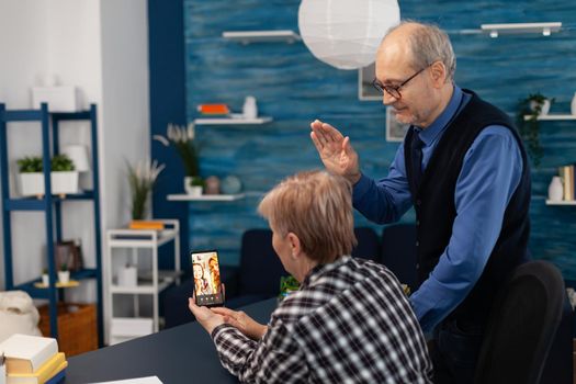 Senior man waving at smartphone camera during video call with relatives. Happy grandparents communicating with family via online web internet video conference using modern internet technology while sitting in the living room