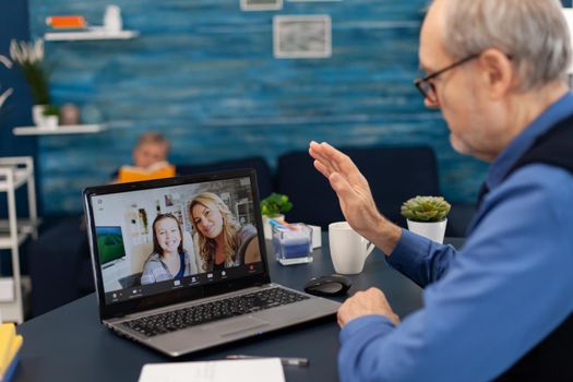 Senior man waving at daughter and niece during video conference. Happy grandfather saying hello in the course of online video confencere with family from living room.