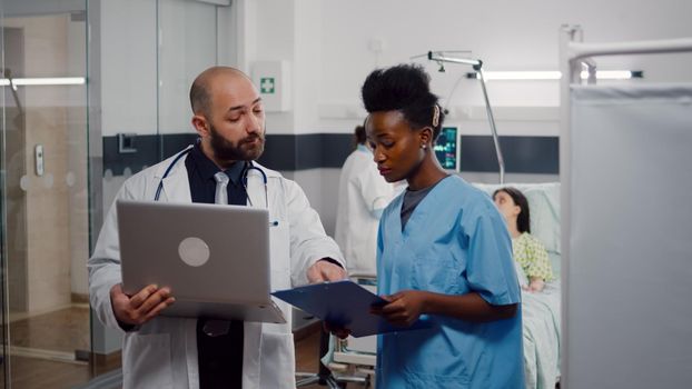 African nurse and surgeon doctor in medical uniform analyzing illness symptom working in hospital ward. In background woman physician checking sick patient while recovering after surgery