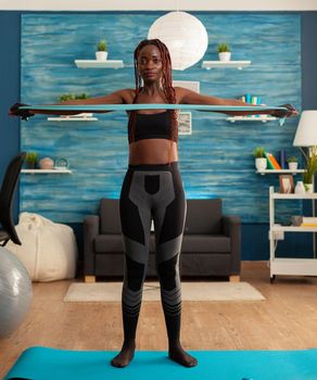 African doing home workout in living room with rubber band on yoga mat for back muscles. Strong fit black woman exercising for healthy body and lifestyle.