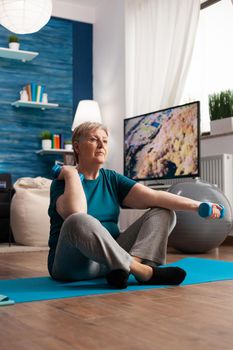 Senior woman in sportwear warming up arm muscles practicing wellness workout using dumbbells. Pensioner sitting on yoga mat in lotus position during aerobics routine looking at gym video