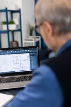 Overshoulder shot of senior architect working on laptop to finish a project. Experienced construction engineer designer looking at house blueprints on laptop computer from home office.