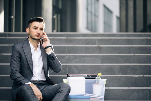 Handsome jobless office worker entrepreneur in formal suit sitting on stairs outdoors. Fired man talking on phone with stuff in box. Cellphone conversation. Speaking on telephone.