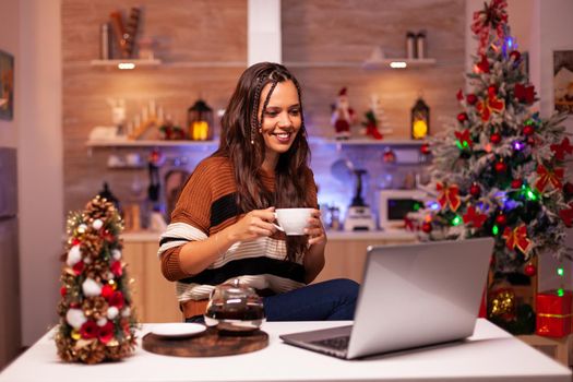 Smiling adult on video call online conference in christmas decorated kitchen at home. Festive young woman talking to friends and family on laptop for seasonal winter celebration
