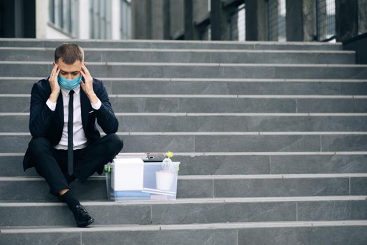 Fired male office worker in medical mask sitting on stairs in depression with box of stuff. Anxious concept. Workless man in despair. Unemployed businessman lost his business.