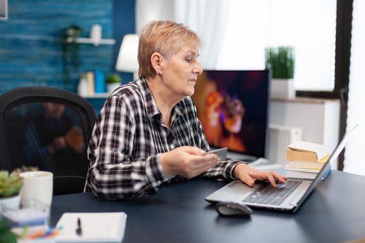 Positive senior woman paying bills holding plastic credit card. Joyful elderly woman using online banking for payment transcation surfing on internet from home living room.