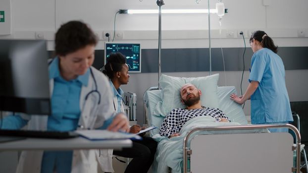 Medical practitioner doctors checking sick man analyzing disease symptom during recovery appointment in hospital ward. Patient resting in bed while assistant arrange to sit comfortable