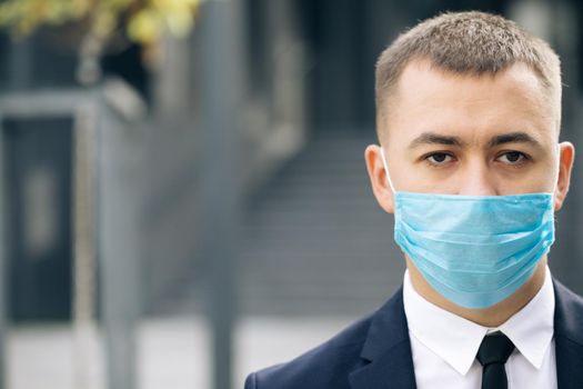 Half face of upset caucasian young handsome man in medical mask looking straight to camera while standing outdoors near modern office building. Man in protection mask look at street.